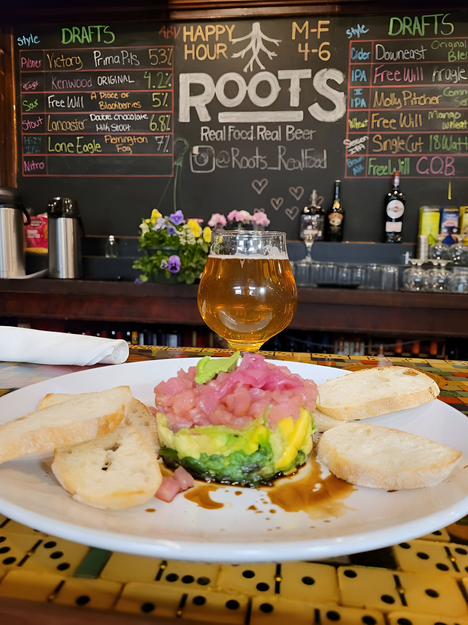 A plate with guacamole and diced onion is garnished with toast slices. A beer glass is placed in the center. A draft beer and food menu is on a chalkboard in the background of a bar area.