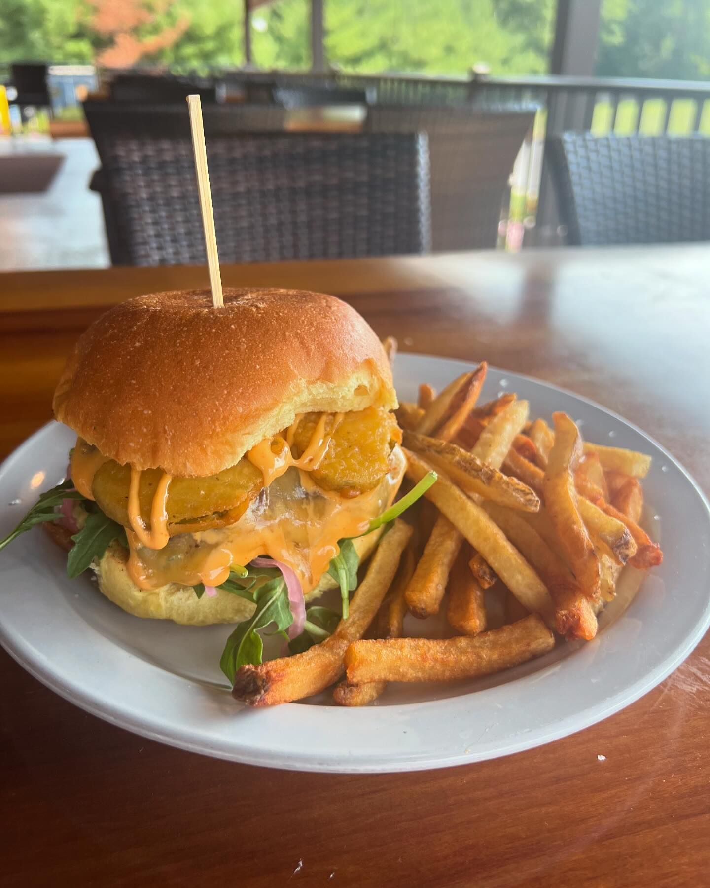 A cheeseburger topped with fried pickles and sauce is served with a side of French fries on a white plate in an outdoor dining setting.