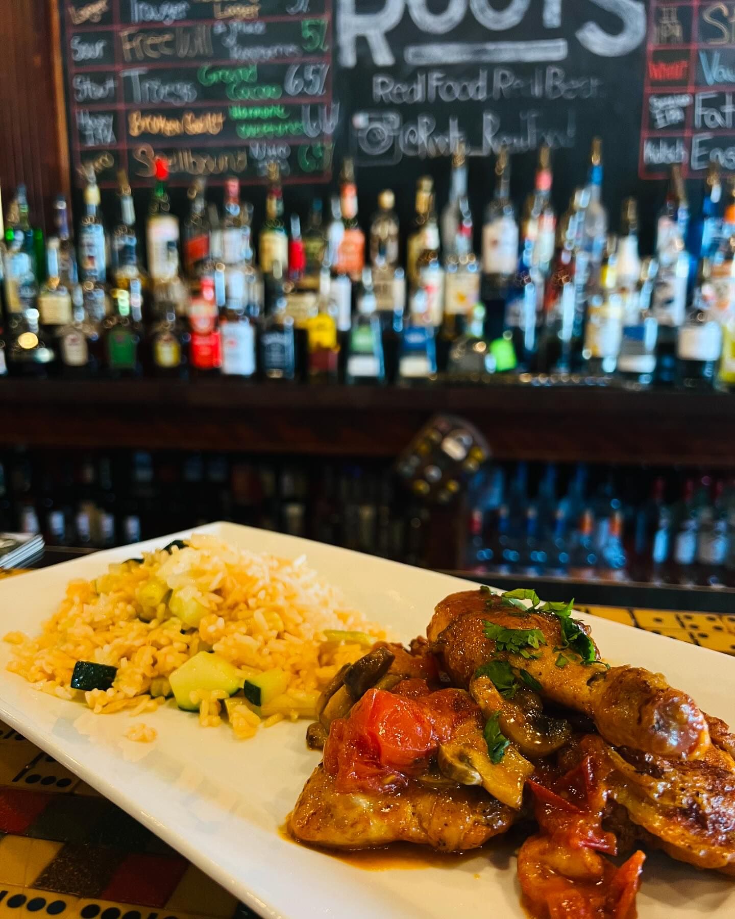 A plate of rice and vegetables with a meat stew is placed on a counter in front of a bar stocked with various bottles. A chalkboard menu is partially visible in the background.