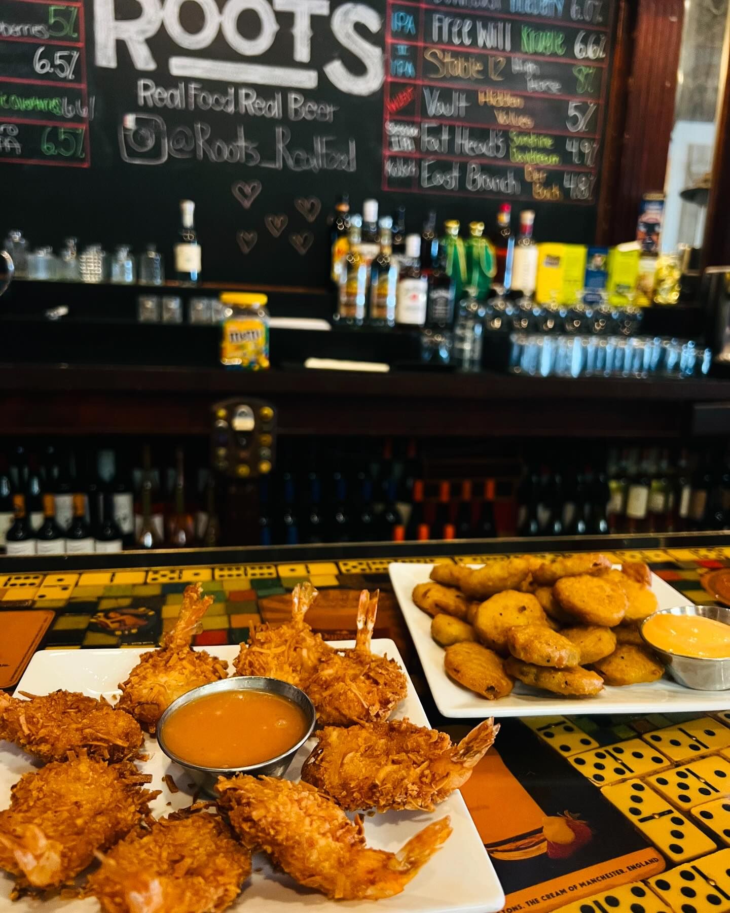 A bar counter with menu boards behind it displays plates of coconut shrimp and patties with dipping sauces in front. In the background, various bottles of alcohol are arranged on shelves.