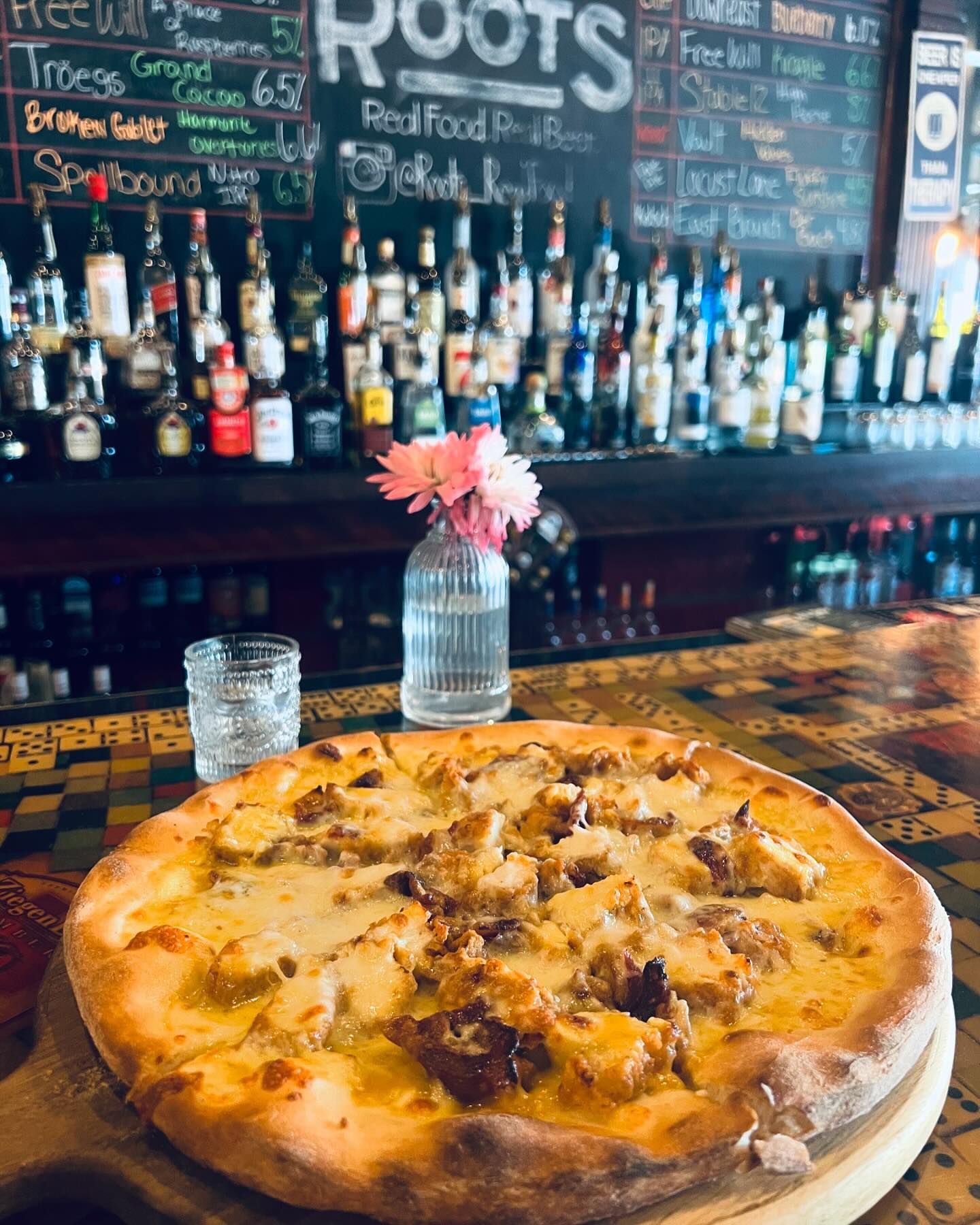 A freshly baked pizza with cheese and chunks of meat is placed on a colorful countertop. Behind it, there is a glass jar with flowers, two glasses of water, and a fully stocked bar with a menu board.