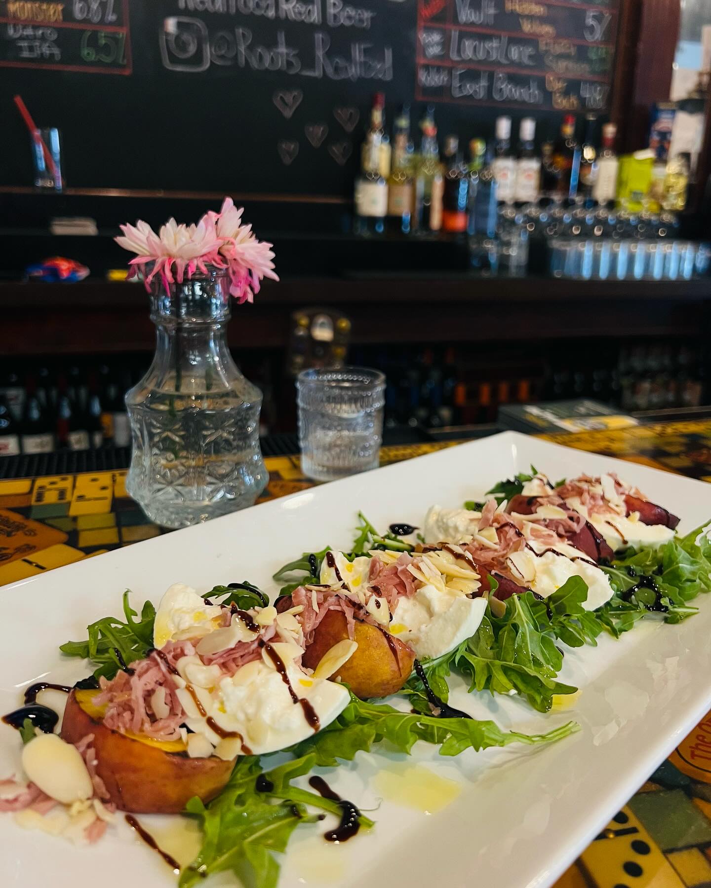 A bar counter with a plate of gourmet appetizers featuring peach slices, arugula, cheese, and drizzle. A glass of water and a vase with pink flowers are also on the counter, with alcohol bottles in the background.