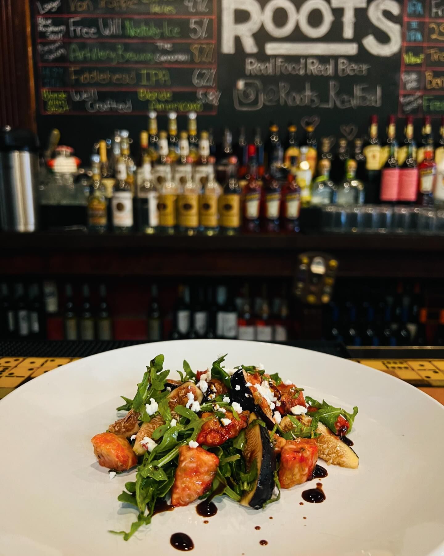A white plate with a fresh salad, featuring arugula, cherry tomatoes, goat cheese, and grilled vegetables, is set on a bar counter with a colorful menu board and bottles in the background.
