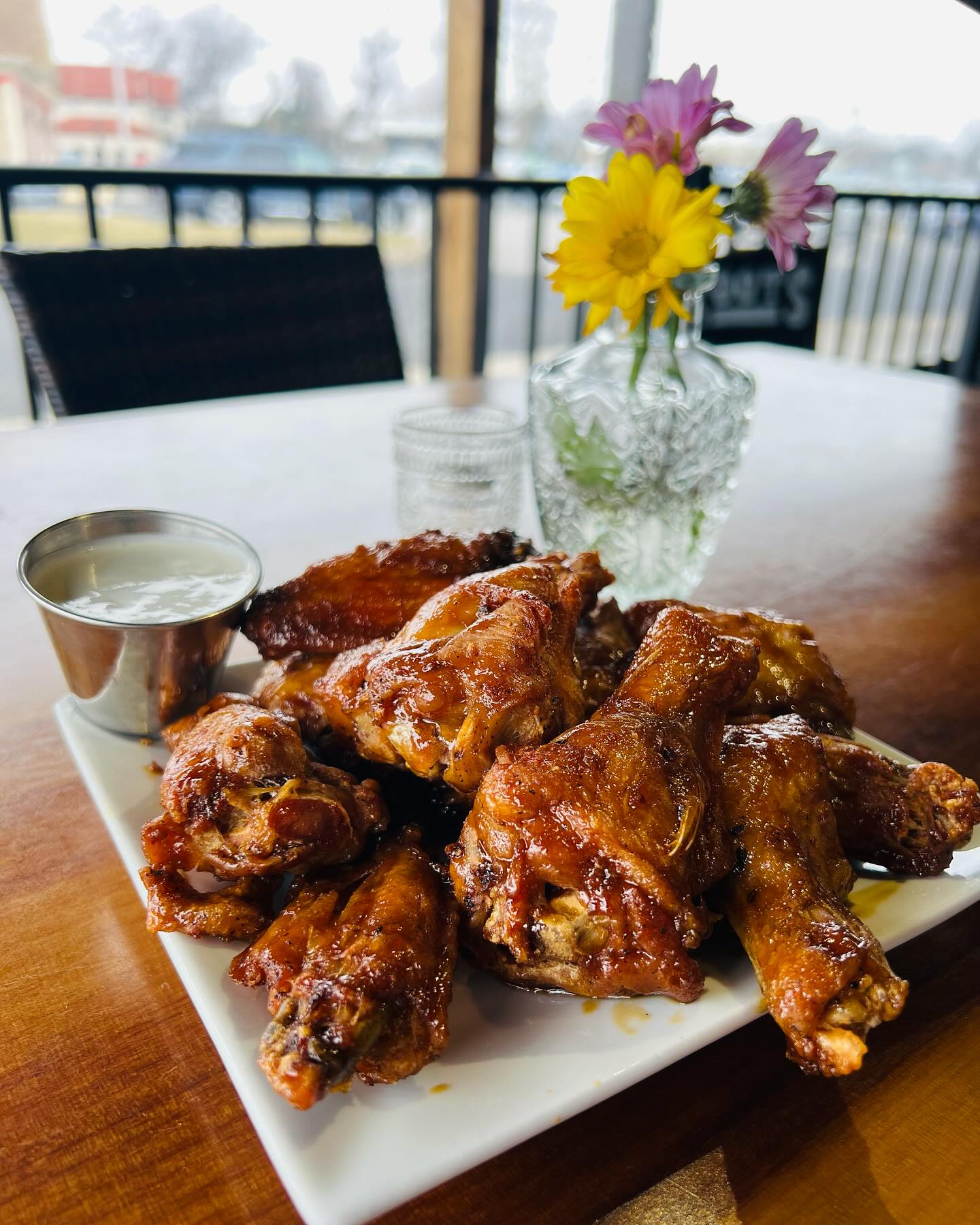 A white square plate of glazed chicken wings is accompanied by a small cup of dipping sauce. A glass vase with yellow and purple flowers is in the background on a wooden table.