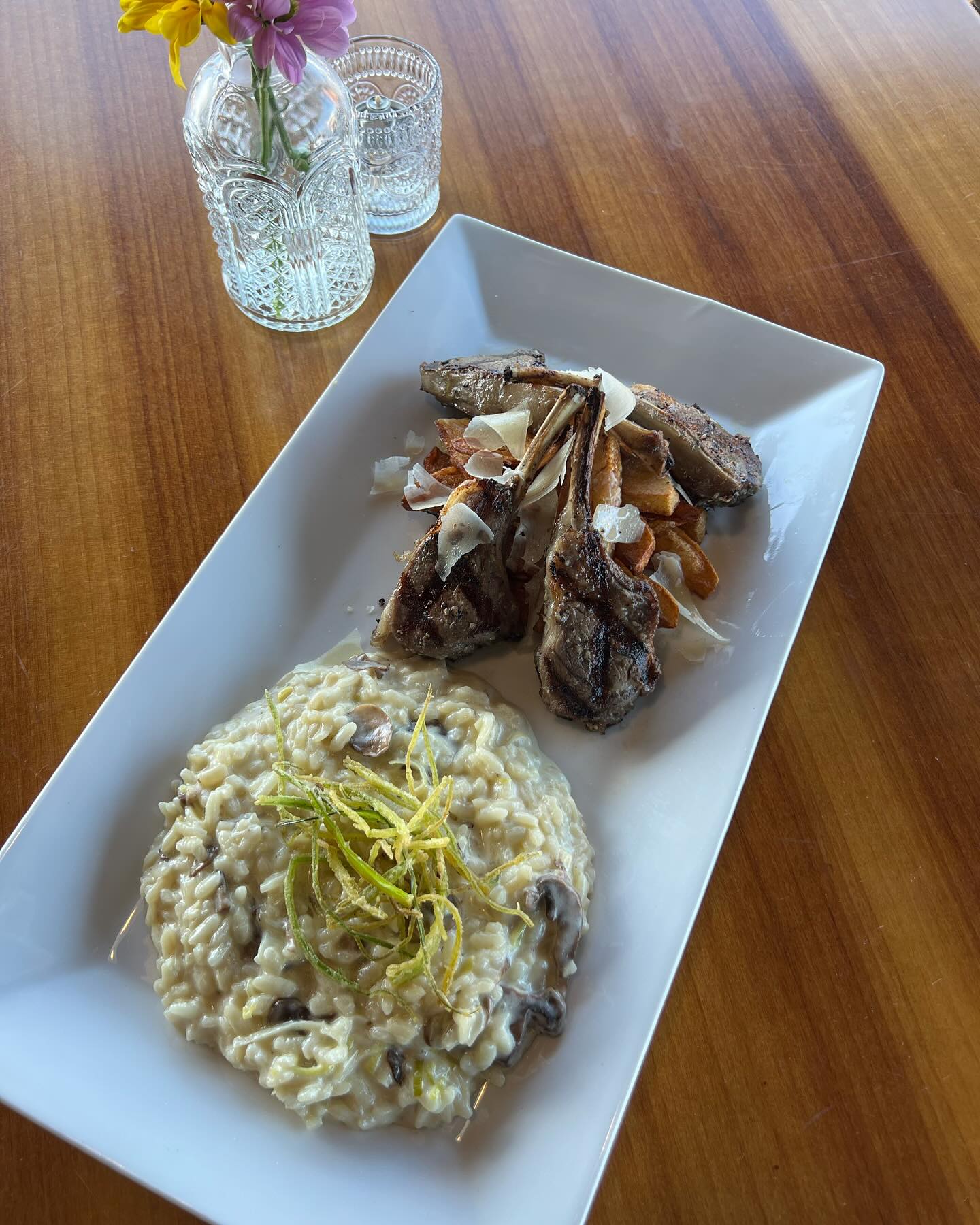 A rectangular white plate with risotto garnished with lemon zest on the bottom left and lamb chops with roasted potatoes on the upper right. The plate is on a wooden table beside two glass vases with flowers.