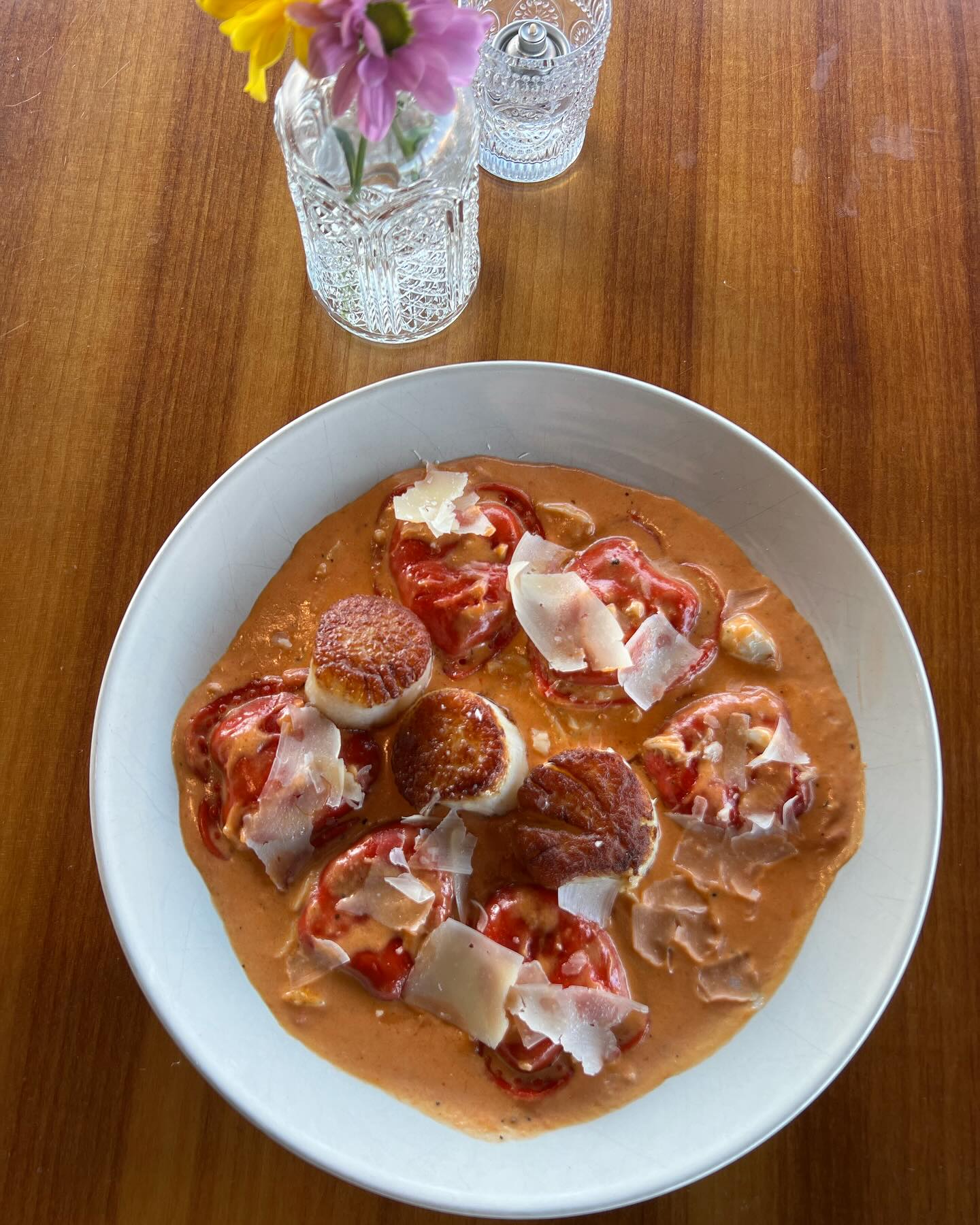 A white plate of tomato-shaped ravioli covered in a creamy sauce, topped with seared scallops and shaved cheese, is placed on a wooden table with a glass vase of flowers in the background.