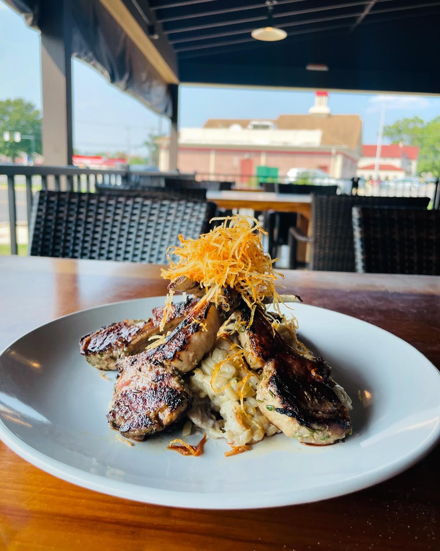 A plate of grilled meat sits atop a bed of creamy risotto garnished with shredded fried vegetables on an outdoor wooden table at a restaurant.
