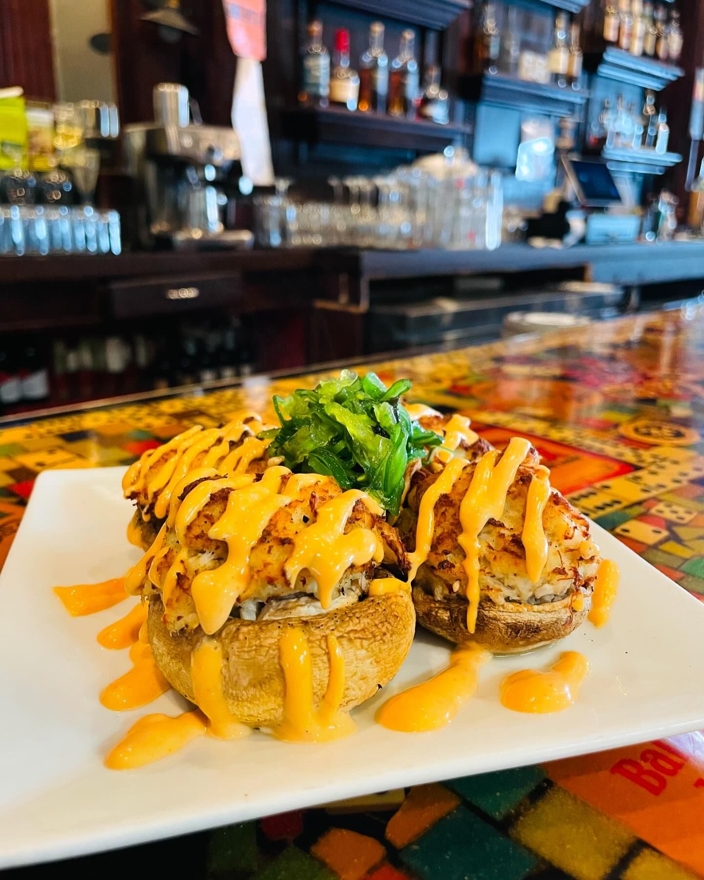 A plate of stuffed mushrooms topped with melted cheese and drizzled with orange sauce, garnished with greens, sits on a colorful, patterned table in a bar setting.