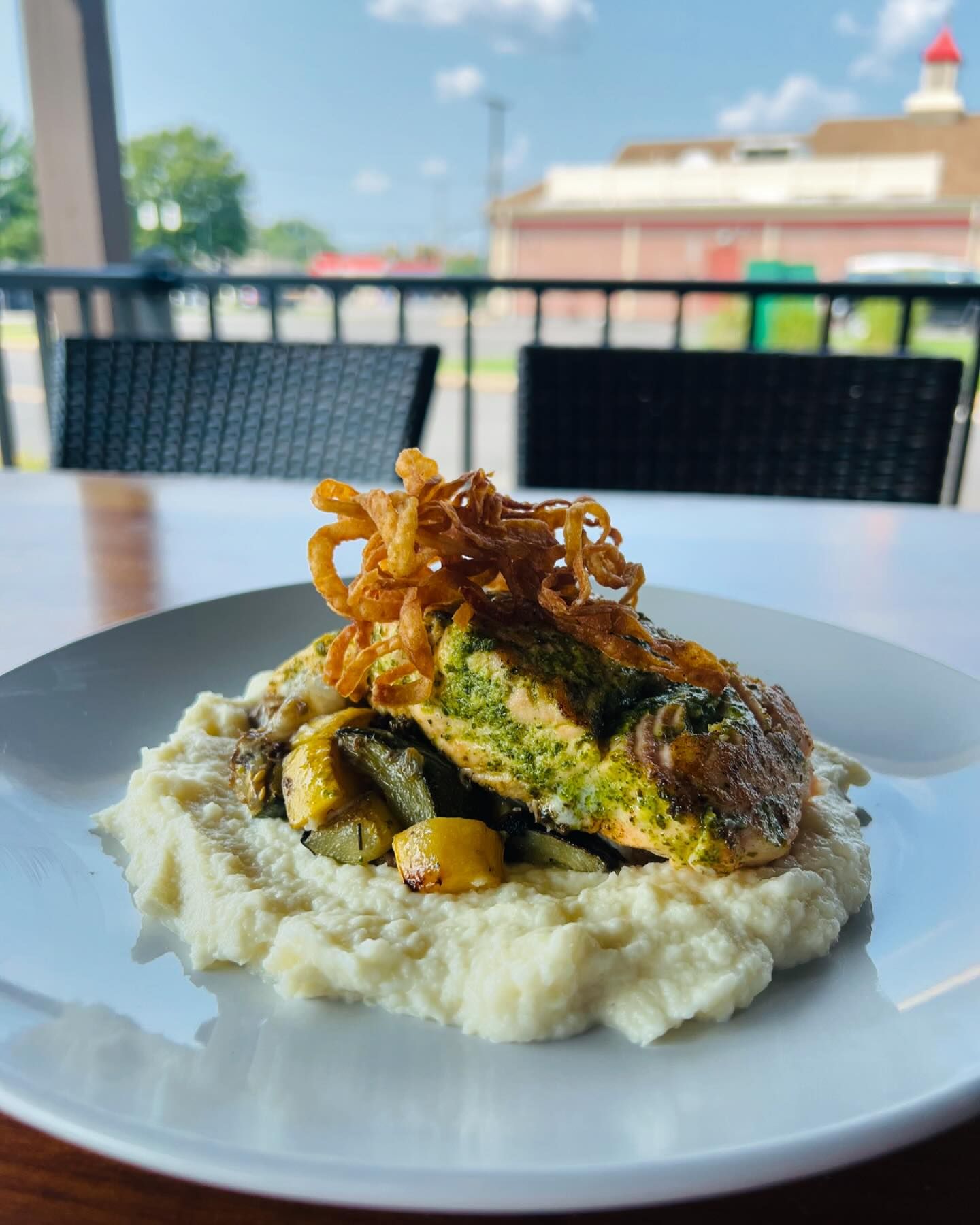 A plate of grilled salmon topped with crispy onion strings, served on a bed of mashed potatoes and roasted vegetables, with outdoor seating and a blurred building in the background.