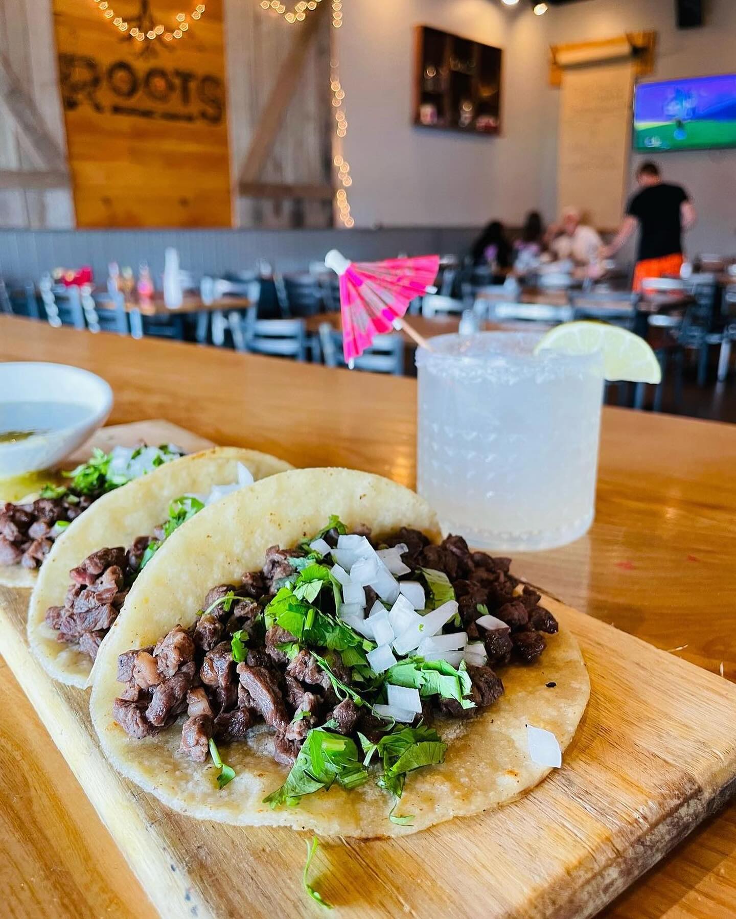 A wooden board with three tacos filled with chopped meat, cilantro, and onions, next to a glass of a clear drink with a lime wedge and a pink umbrella, in a dimly lit restaurant.