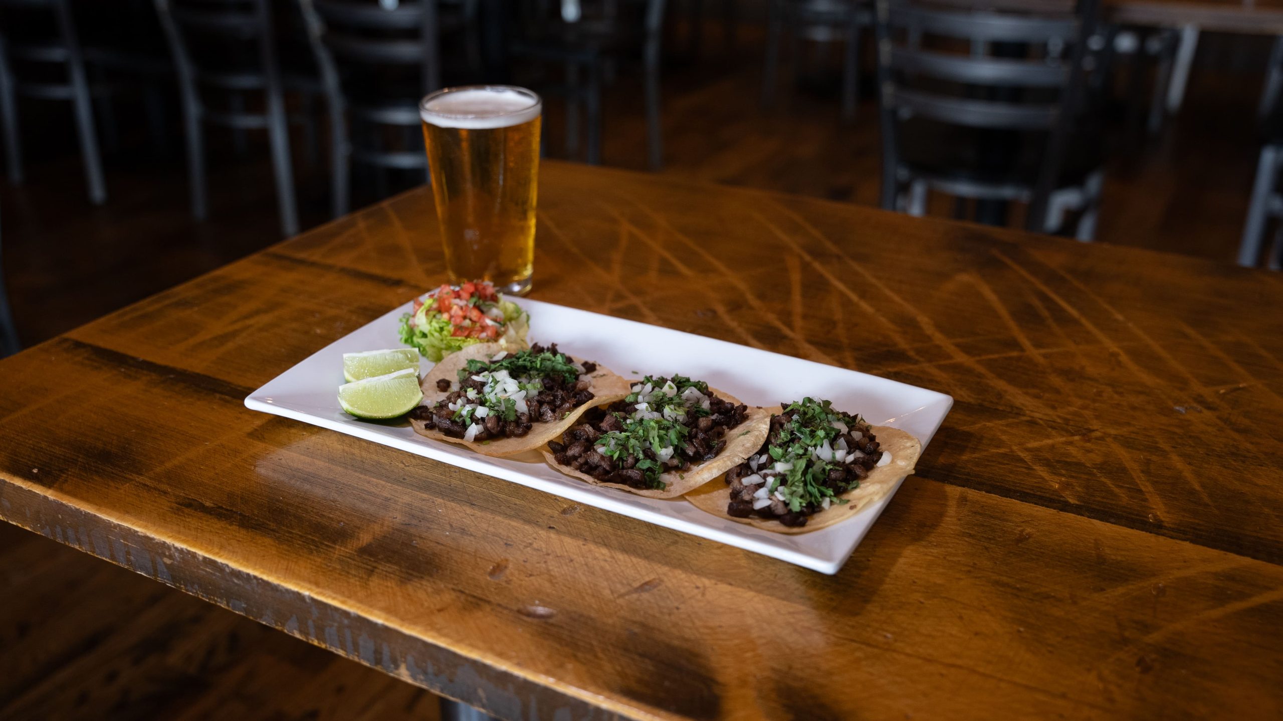 A plate of three tacos with meat, onions, and cilantro, accompanied by lime wedges and salsa, sits on a wooden table. A glass of beer is next to the plate.