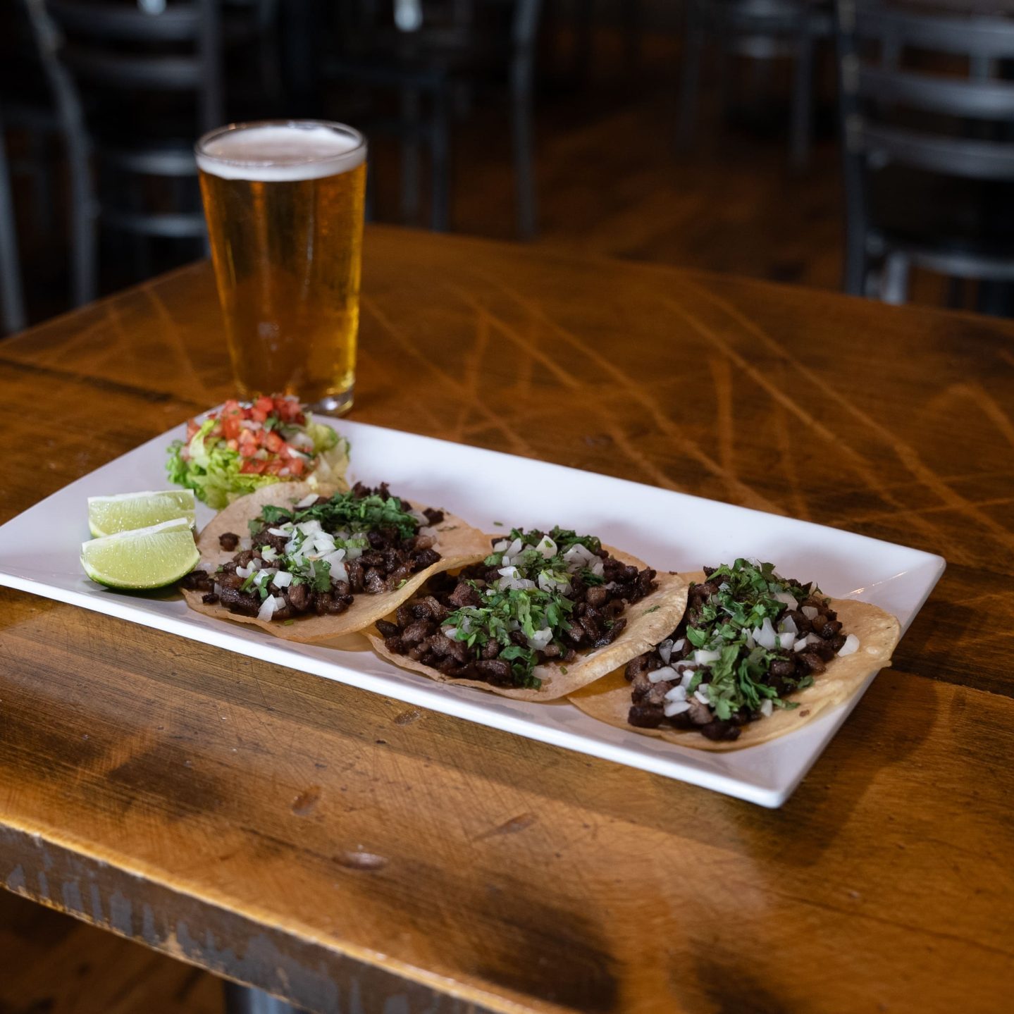 A plate of three tacos with meat, onions, and cilantro, accompanied by lime wedges and salsa, sits on a wooden table. A glass of beer is next to the plate.