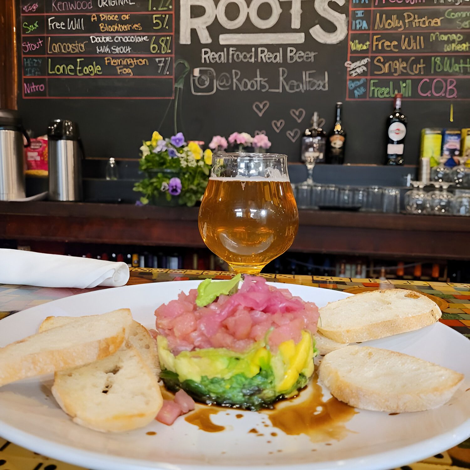 A plate with guacamole and diced onion is garnished with toast slices. A beer glass is placed in the center. A draft beer and food menu is on a chalkboard in the background of a bar area.