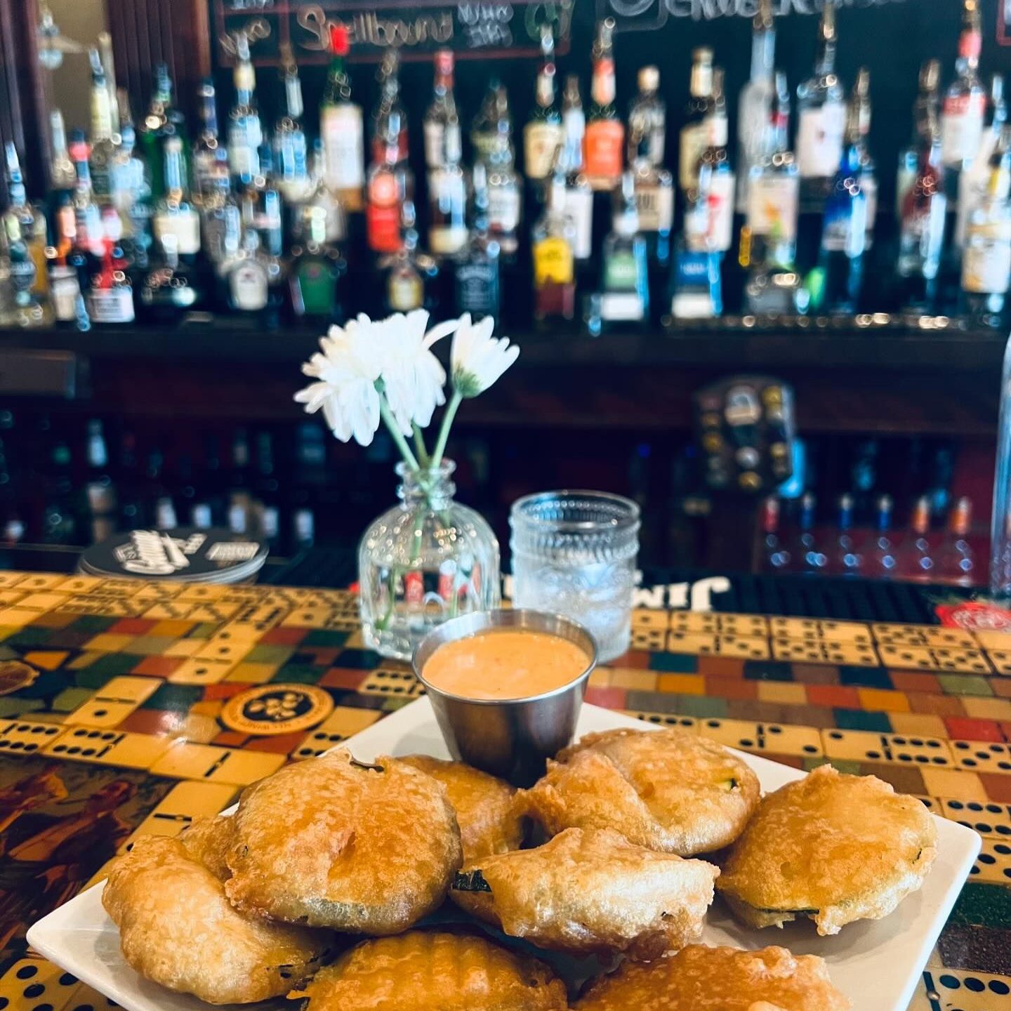 A plate of fried appetizers with dipping sauce on a bar counter, featuring a glass vase with white flowers in the background and a variety of liquor bottles on display.